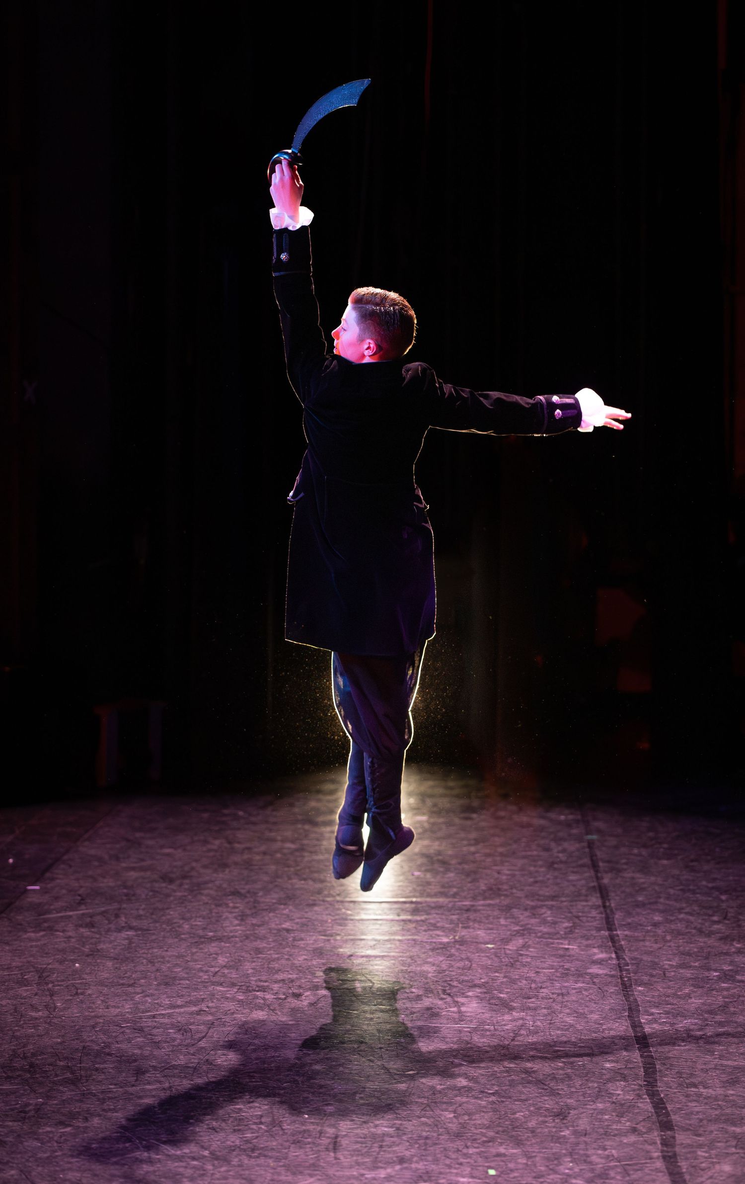 A silhouetted performer leaps dramatically on a dark stage with arms outstretched under spotlights.