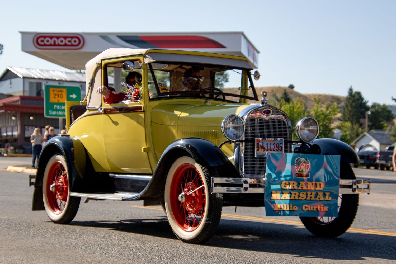 Vintage yellow Ford Model A coupe parked at a gas station with red wheels and classic styling details.