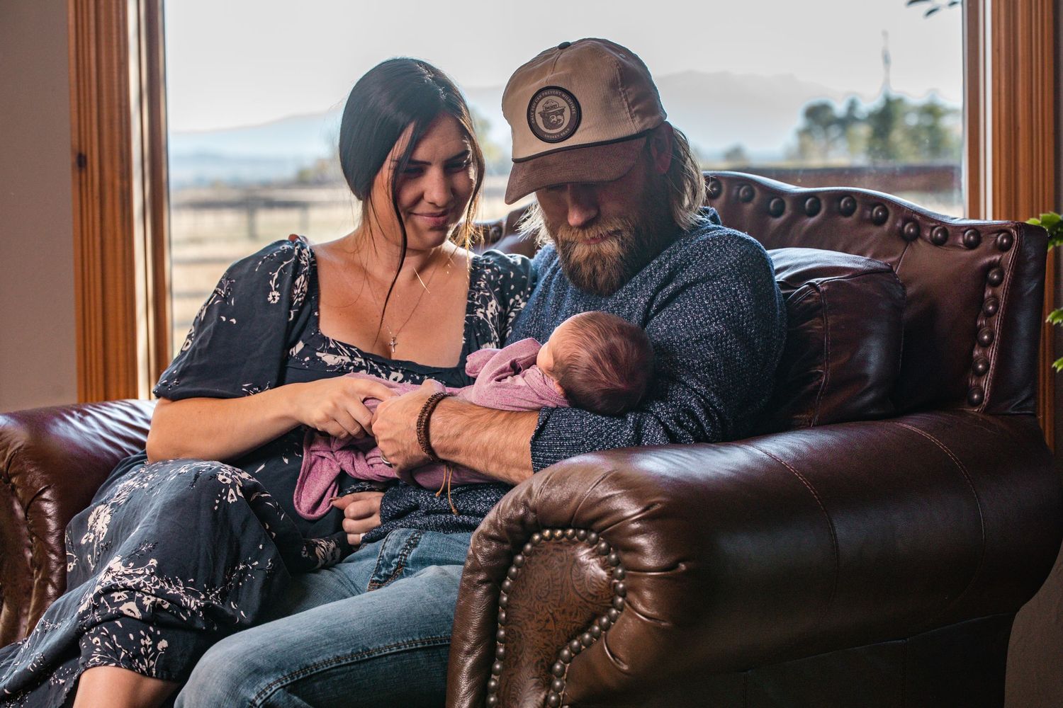 A couple sits together on a brown leather couch while holding a sleeping newborn baby.