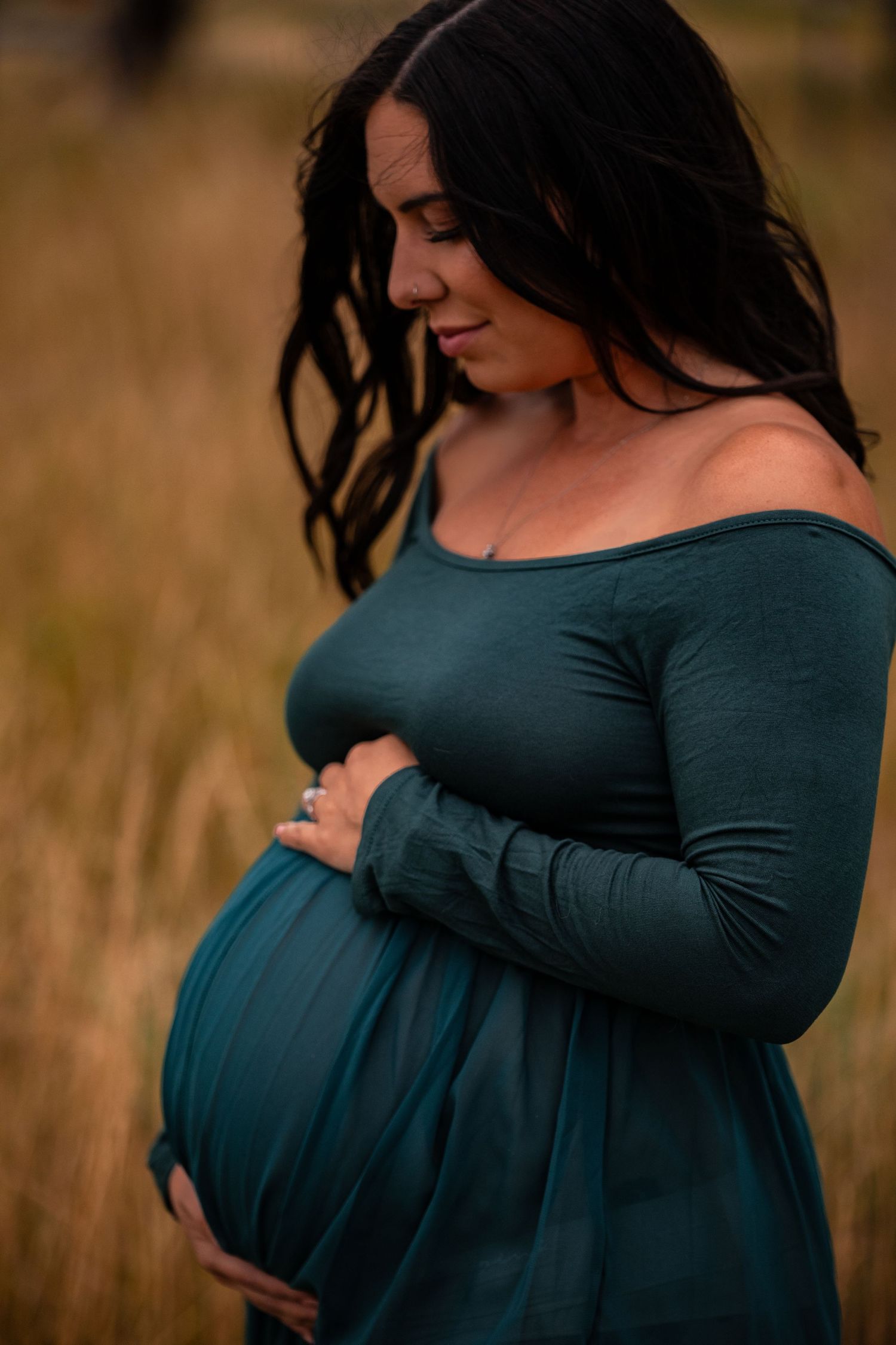 A pregnant woman in a dark teal dress cradles her belly while standing in a golden field at sunset.