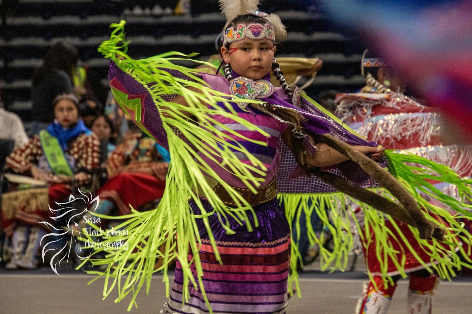 Traditional dancer wearing bright neon green fringe regalia performing at indoor powwow celebration.