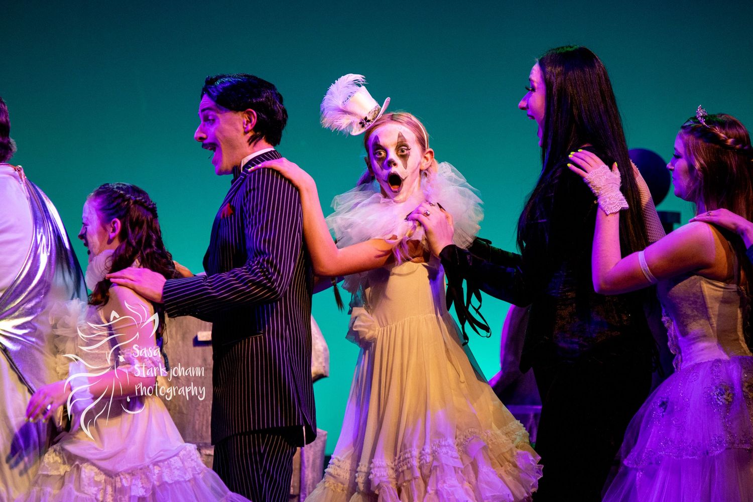 Dancers in formal attire performing on stage with teal lighting during theatrical production.