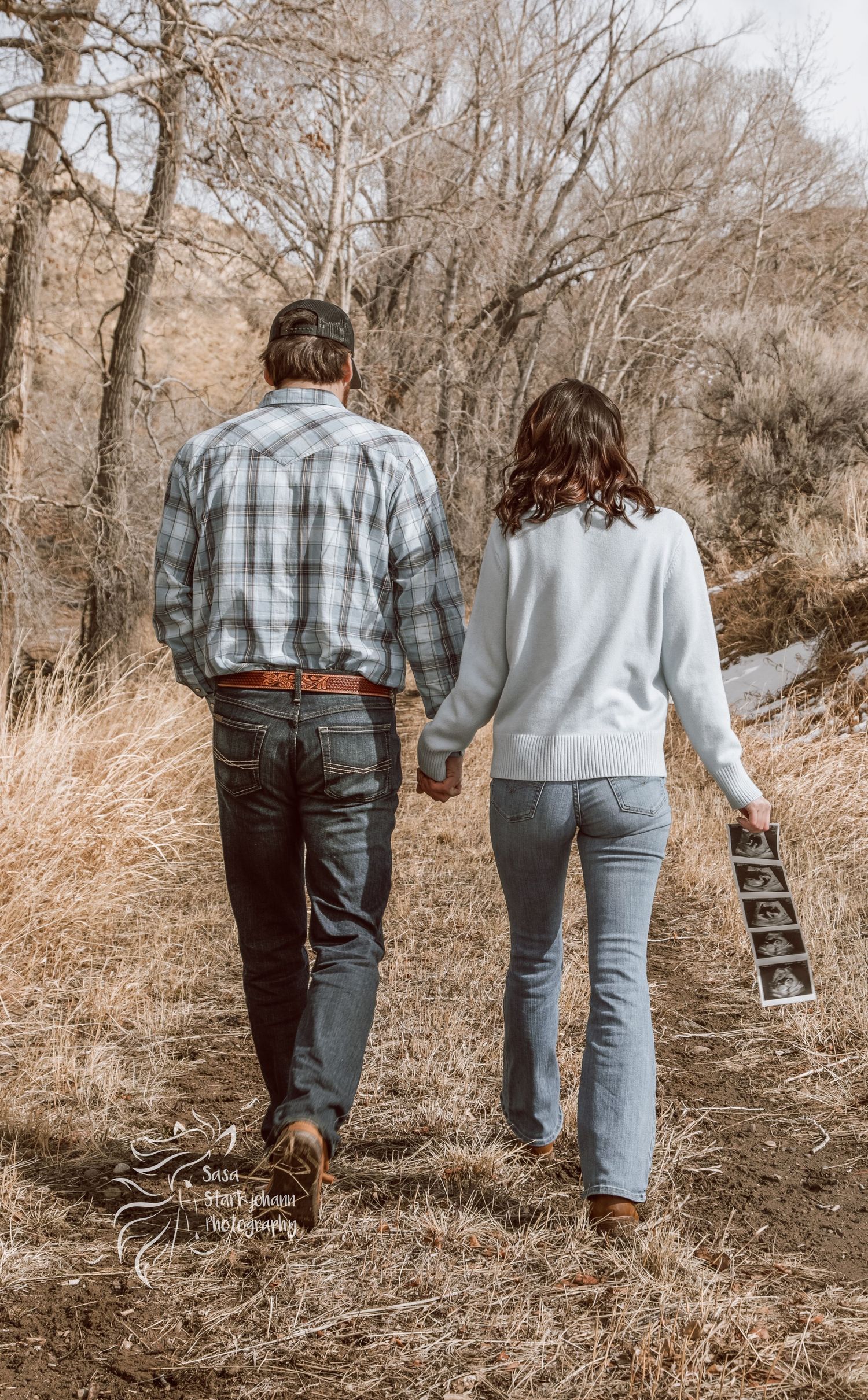 Couple walking away holding ultrasound photos on nature trail.