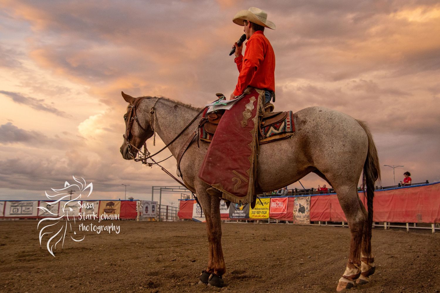 Cowboy in red western wear riding white horse during sunset at rodeo grounds.