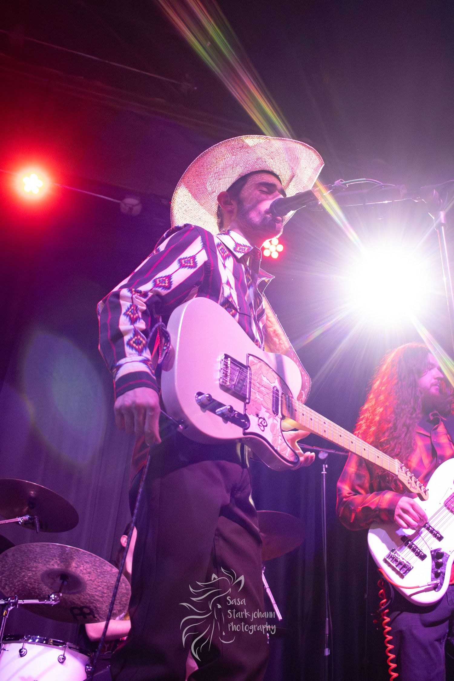 A performer in a cowboy hat plays guitar under bright stage lights during a live show.
