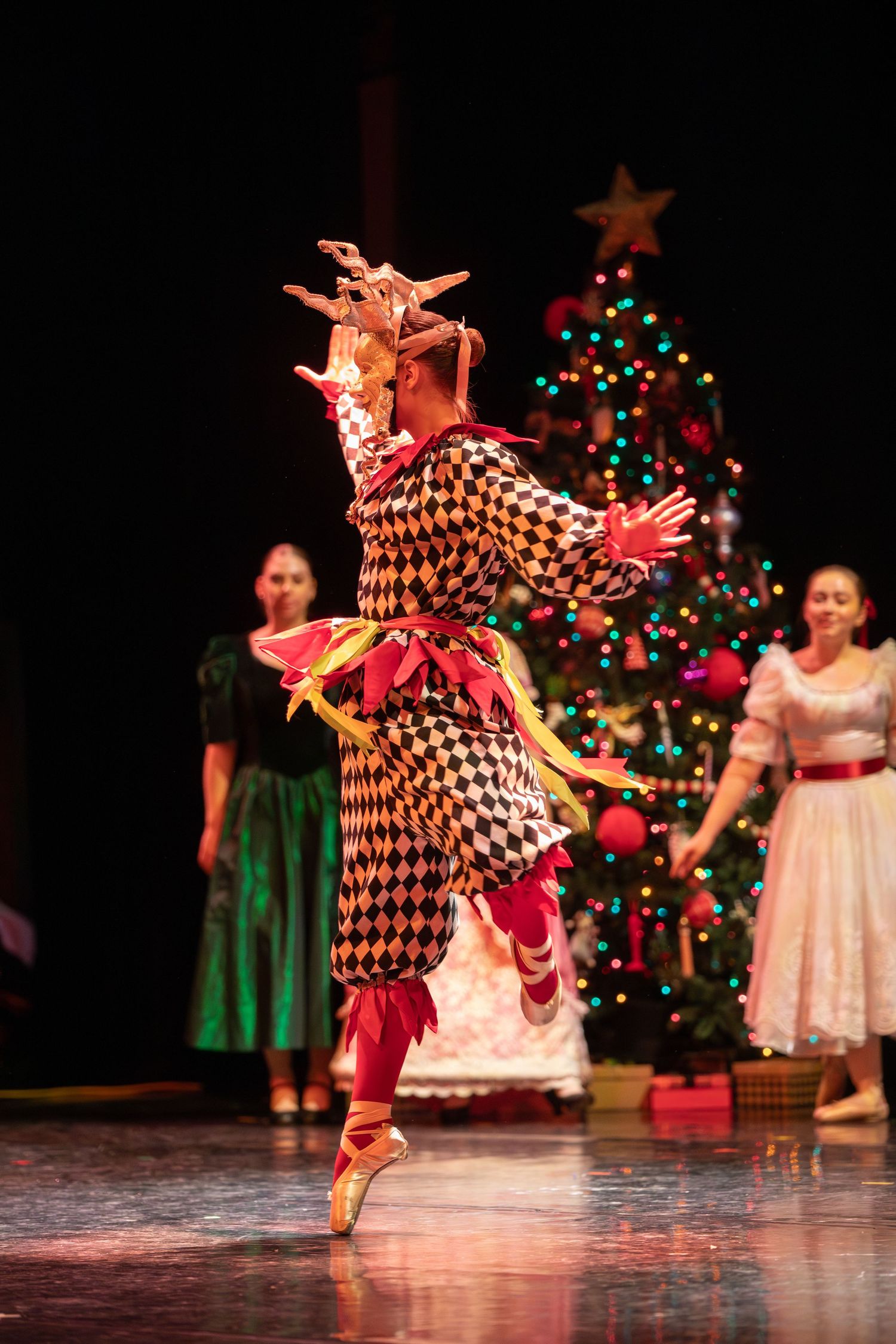 Performer in harlequin costume dances en pointe in front of Christmas tree.