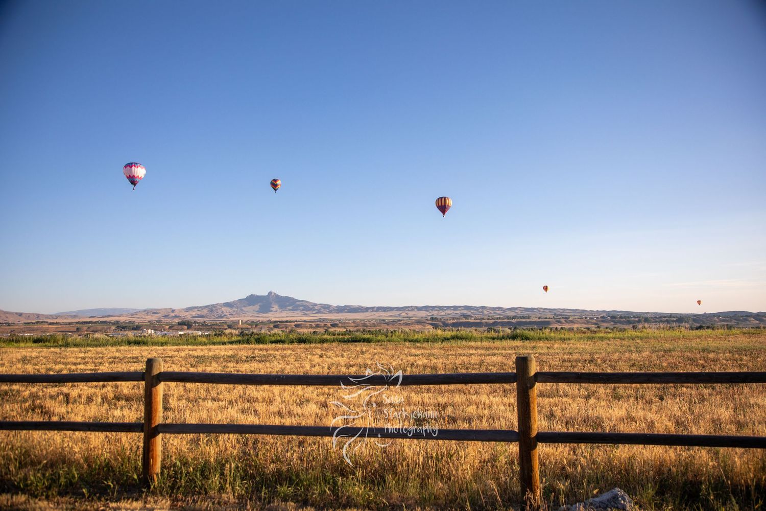 Hot air balloons float over a vast desert landscape with mountains in the distance and wooden fence posts in the foreground.