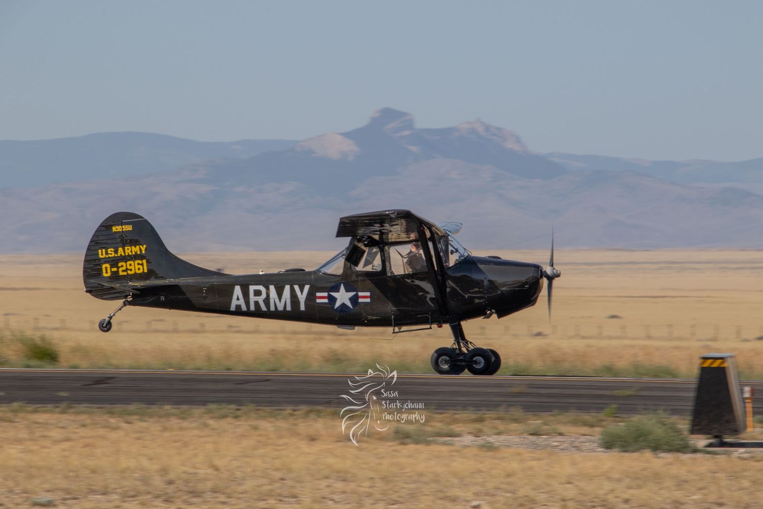 Black US Army L-4 Grasshopper aircraft flying low over desert terrain with mountains in background.