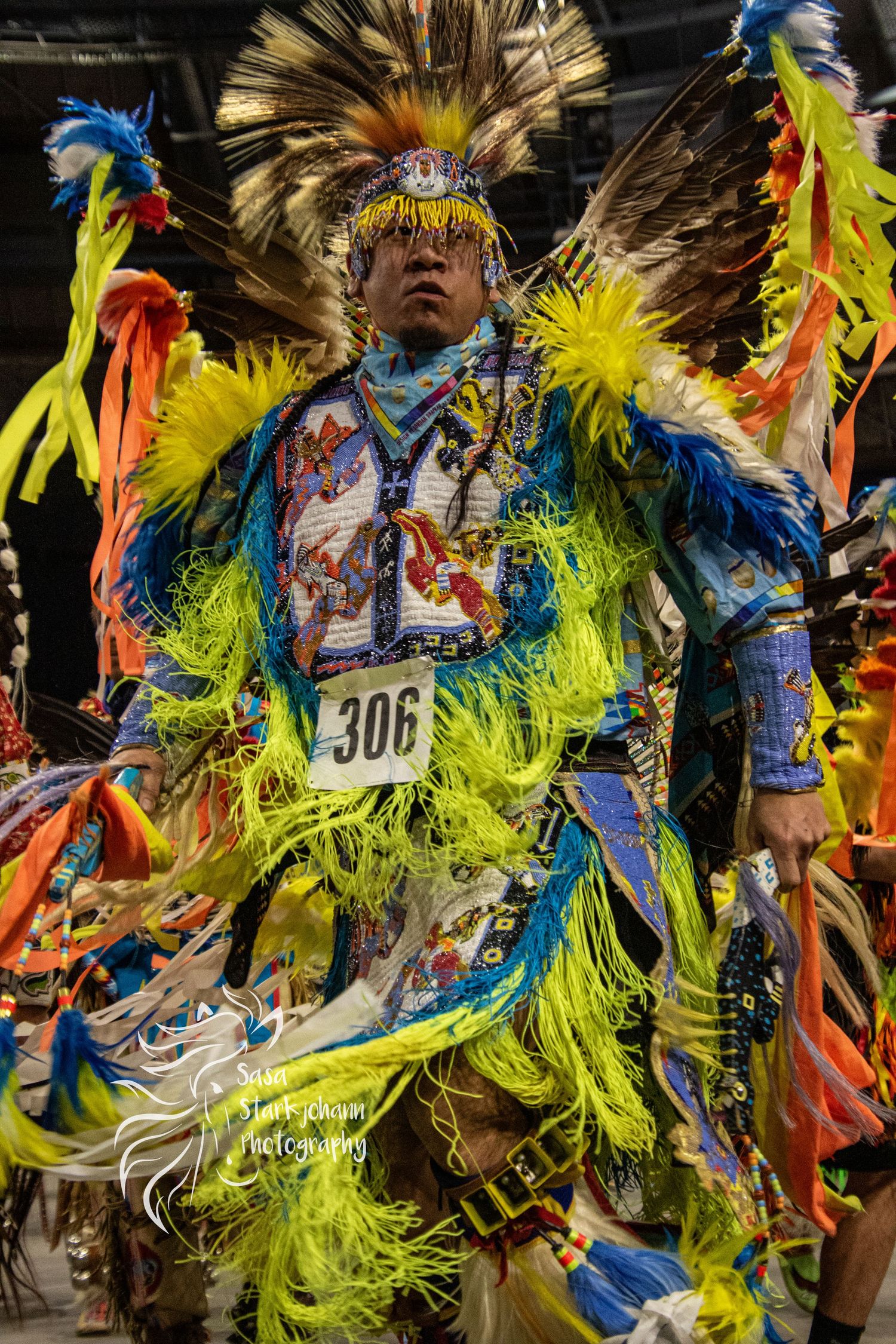 Powwow dancer in vibrant yellow and blue regalia with feathered headdress performing.
