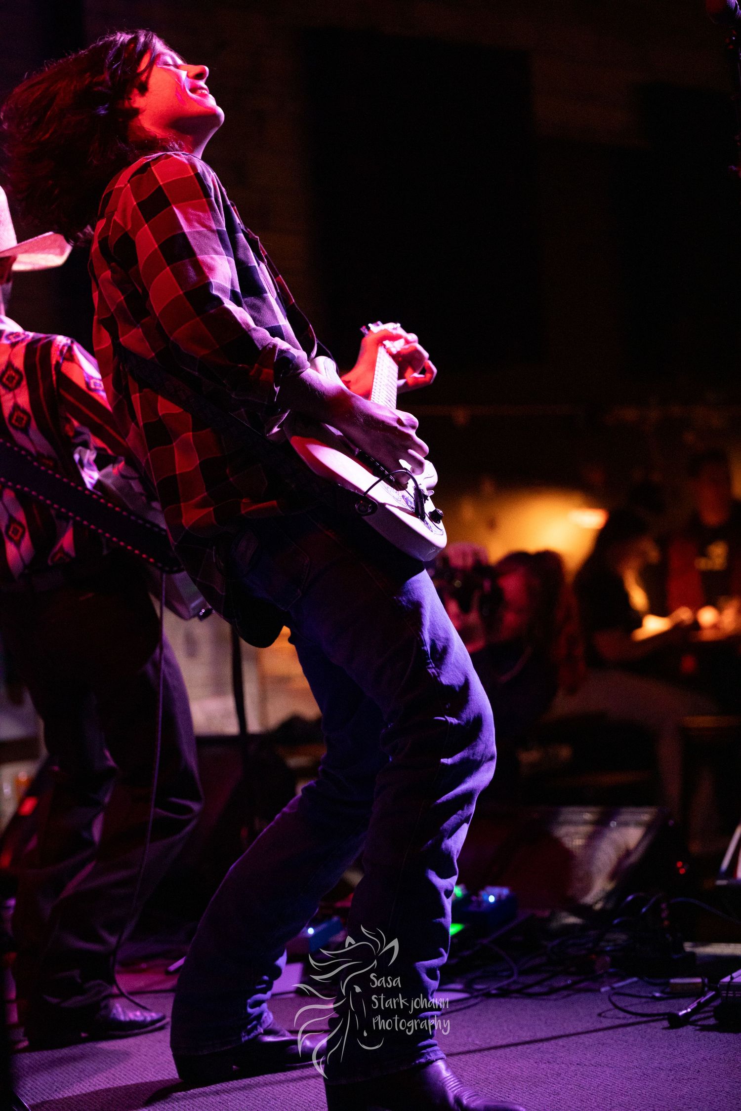 Musician in red plaid shirt performs energetically on stage under dramatic concert lighting.