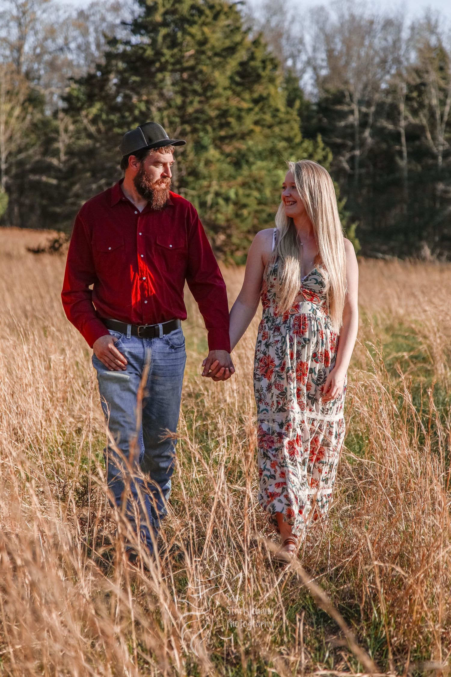 A couple walks hand in hand through tall golden grass at sunset.