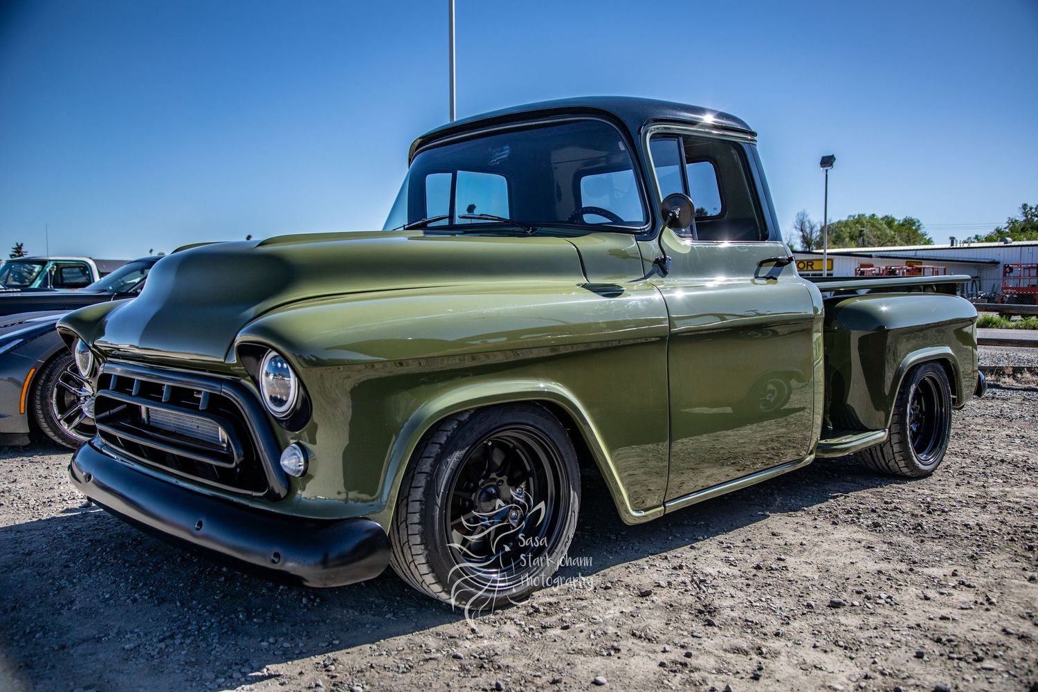 A restored vintage Chevrolet pickup truck in olive green with custom black wheels parked on gravel.