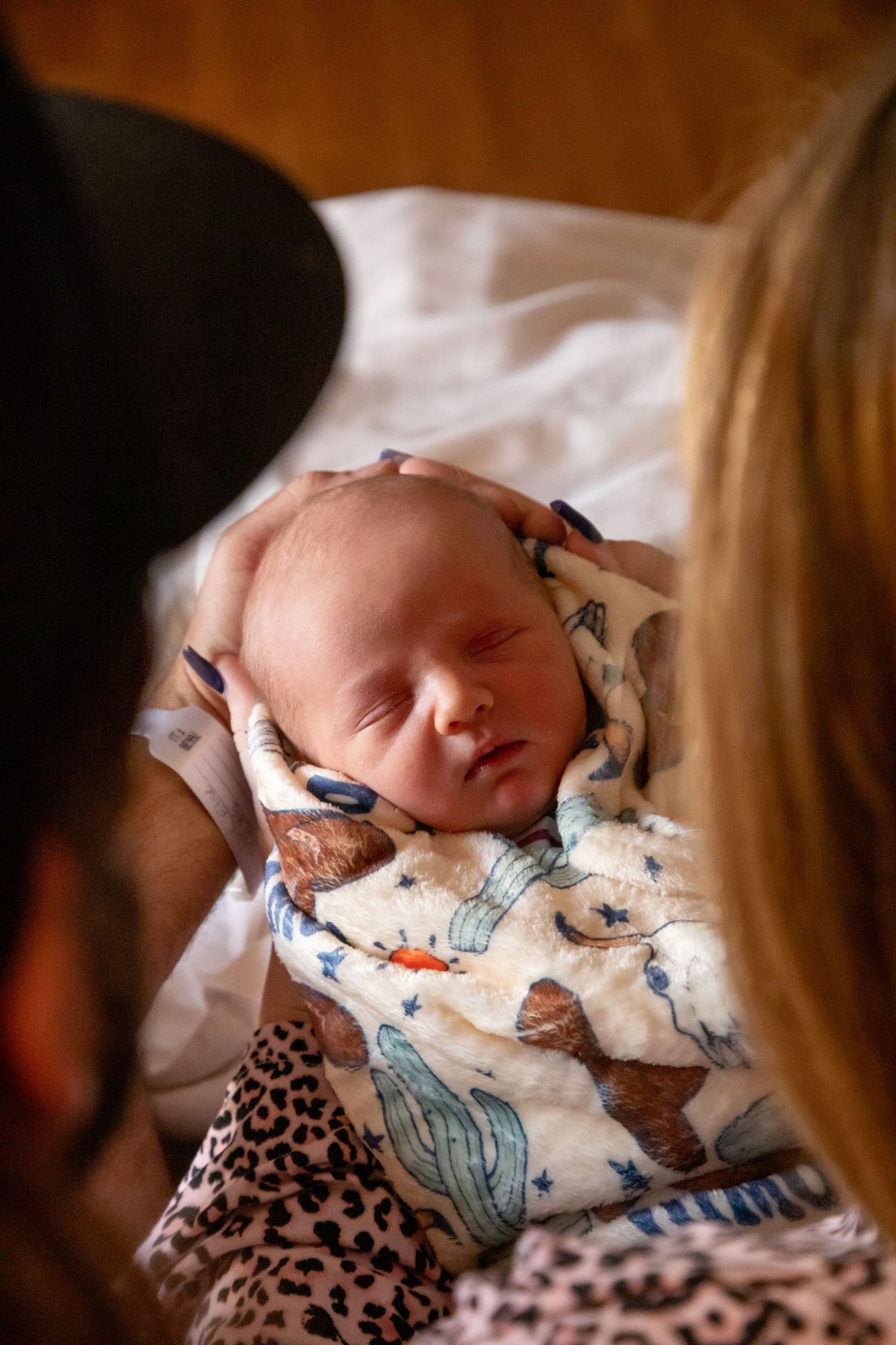Sleeping newborn baby wearing patterned onesie rests peacefully on white bedding.