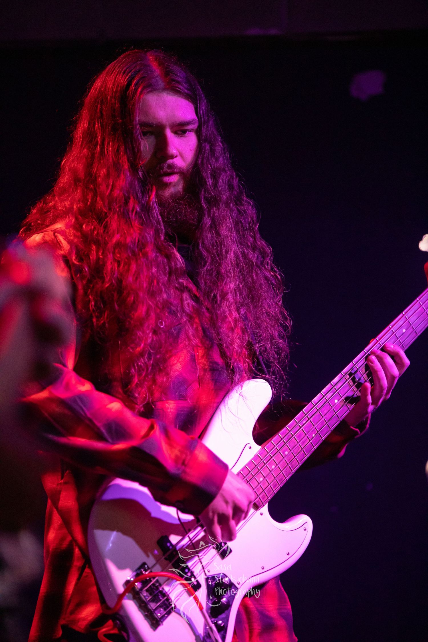 Guitarist with long hair plays white bass under red stage lighting.