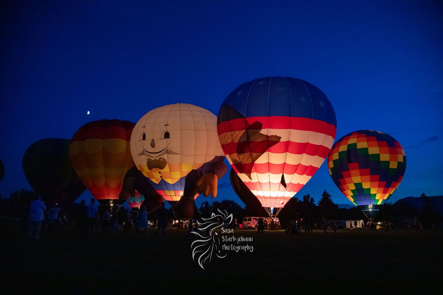 Hot air balloons glowing at dusk during a nighttime balloon glow event against a dark blue sky.