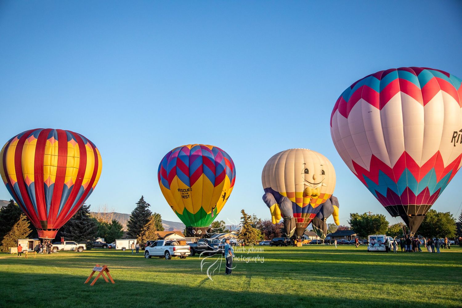 Colorful hot air balloons preparing for launch at sunrise across a grassy field during a festival gathering.