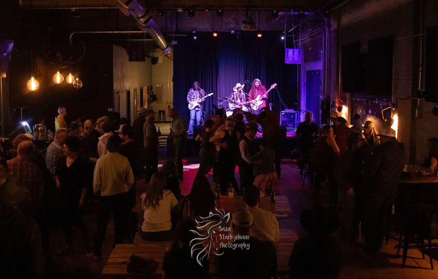 A crowded music venue with audience watching performers on a lit stage in the background.