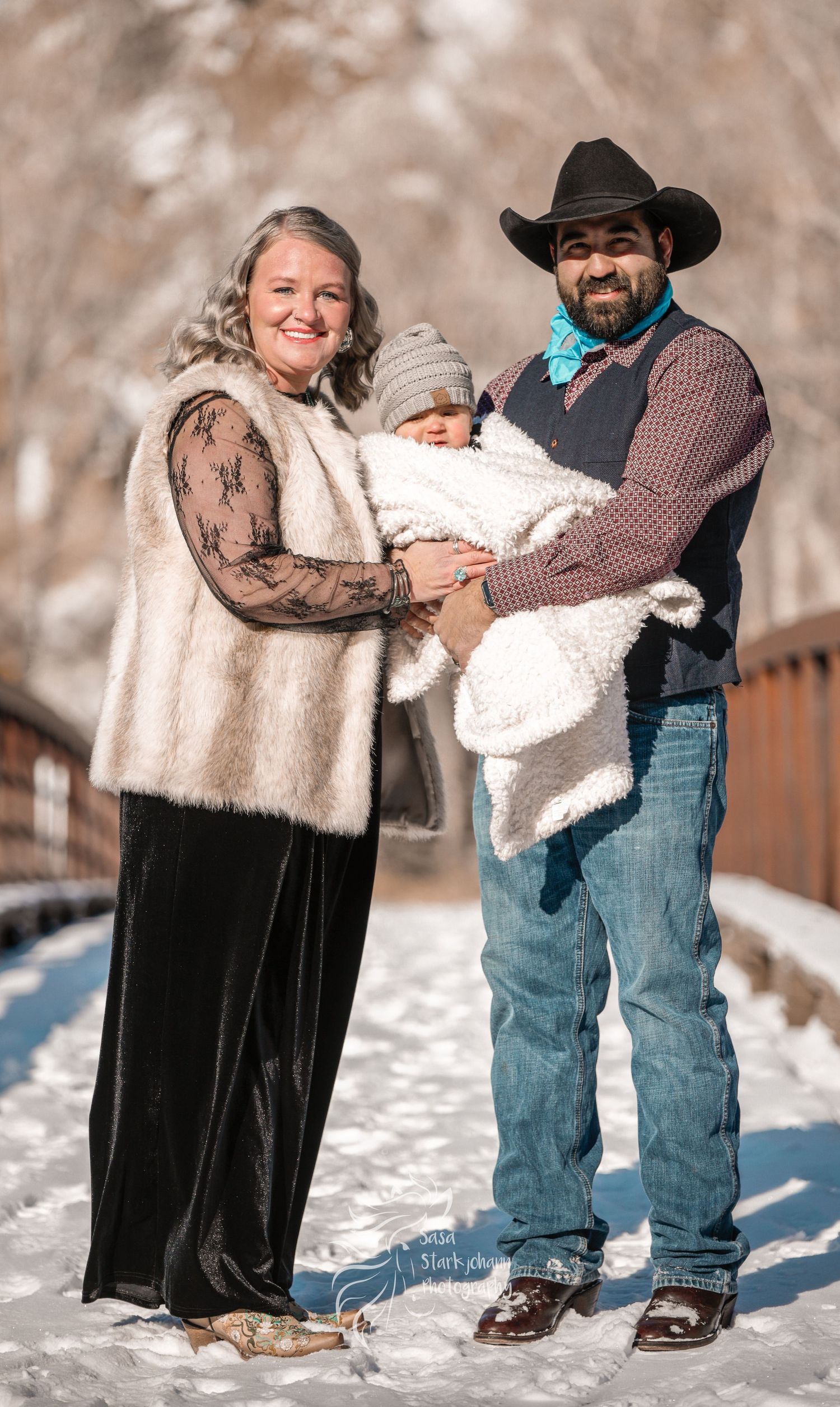 Family portrait of parents with baby standing in snowy winter setting.