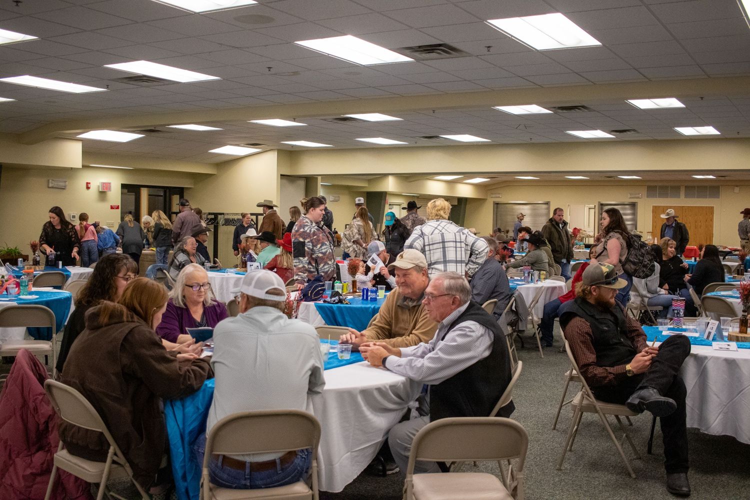 Crowded dining hall with people seated at round tables covered in blue tablecloths.