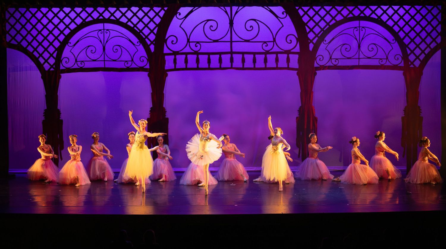 Group of ballet dancers in white tutus perform synchronized choreography under ornate purple archways.