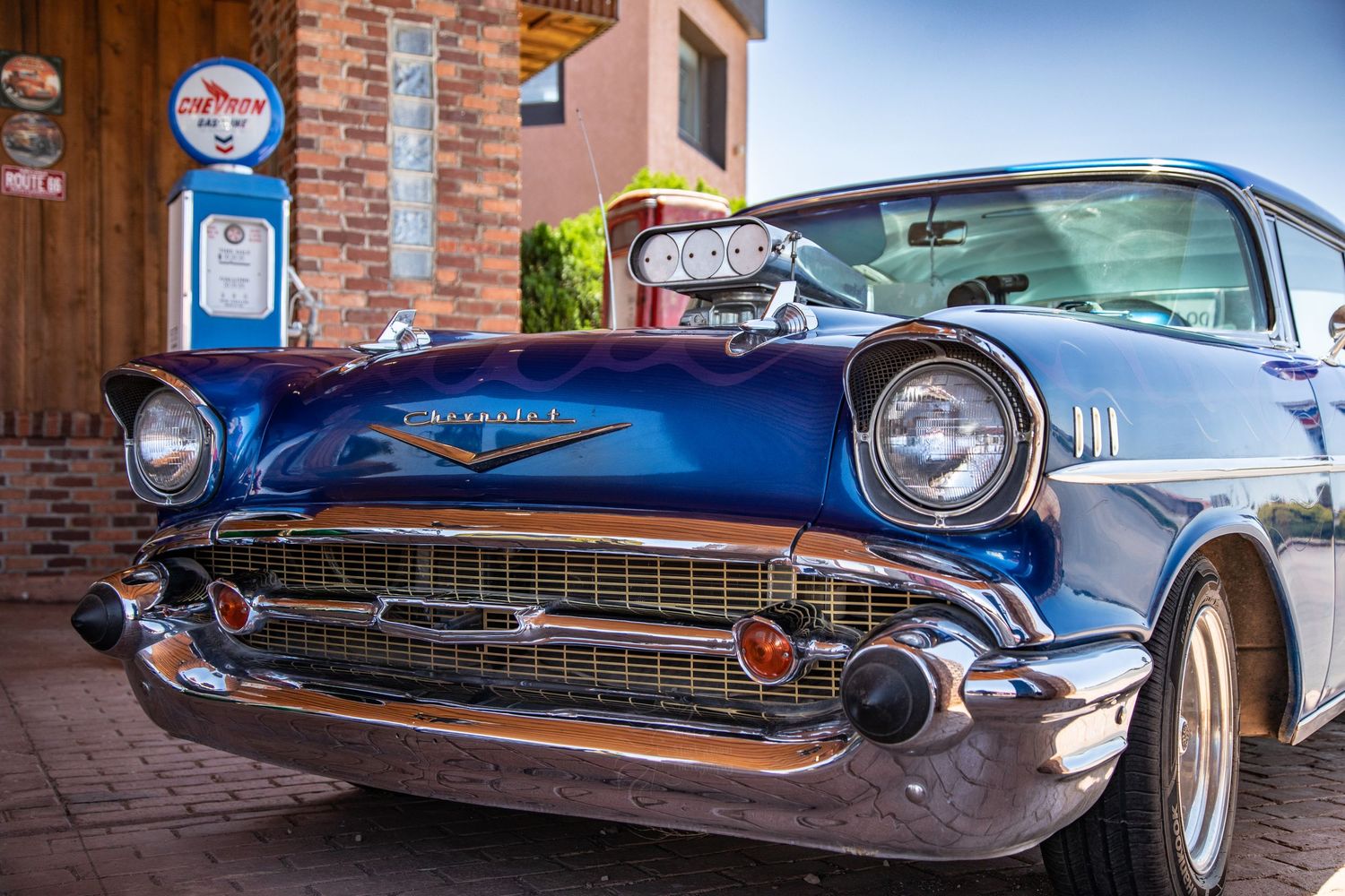 Classic blue 1957 Chevrolet Bel Air with chrome grill parked next to vintage gas pump at brick building.