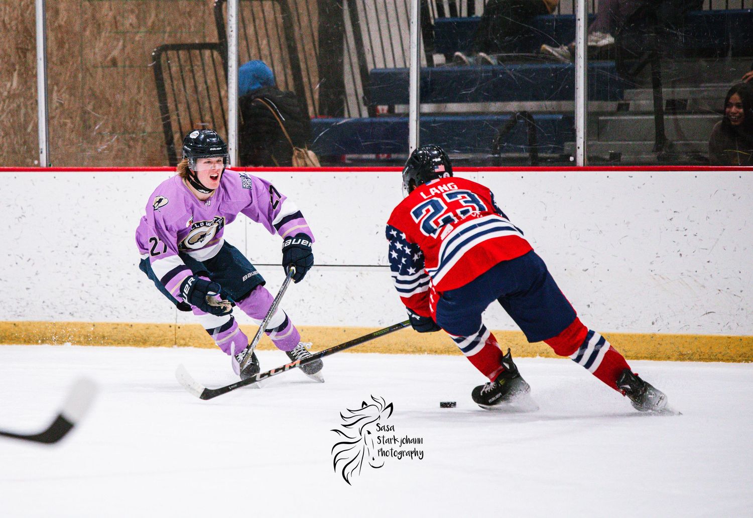 Two hockey players battle for the puck during a game, one in pink and one in red and blue jerseys.