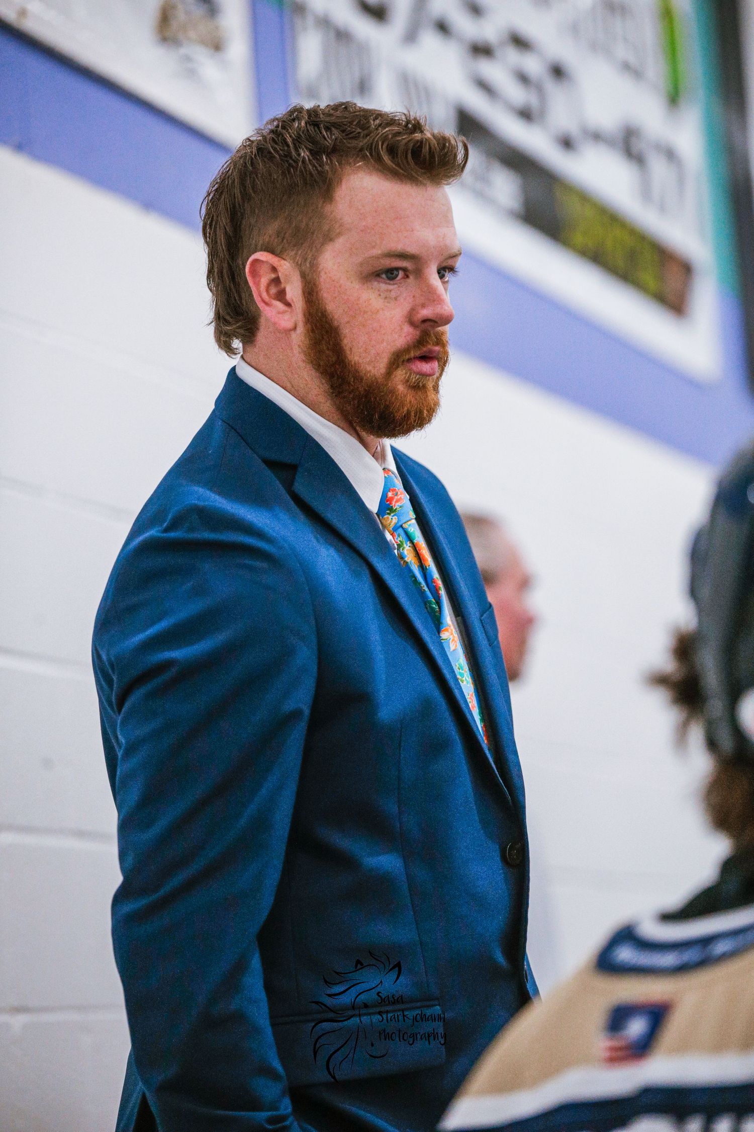 Coach in blue suit watching hockey game from behind bench area.