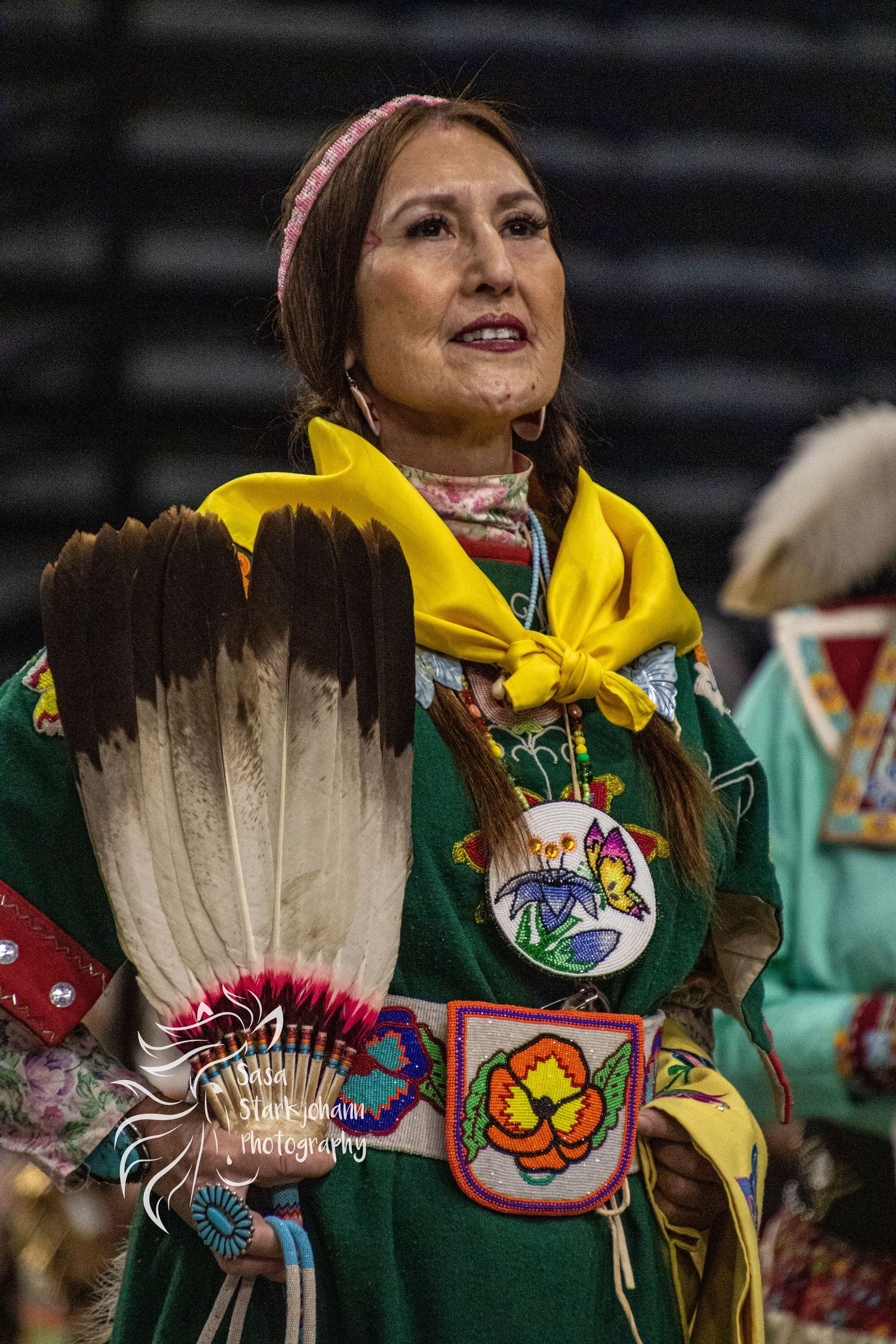Dancer wearing green traditional dress with eagle feather fan and yellow scarf at powwow gathering.