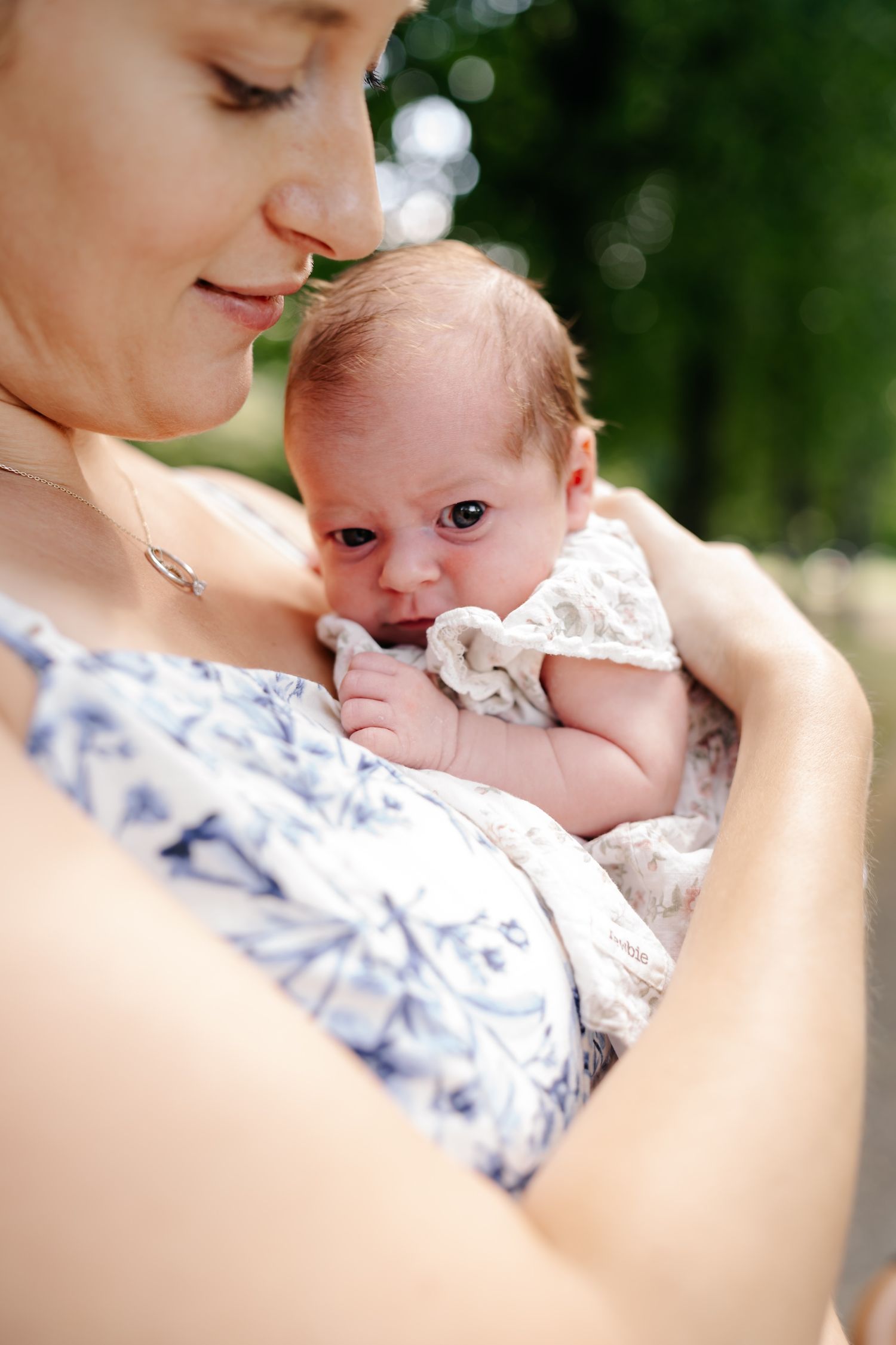 A mother tenderly holds her newborn baby outdoors, both dressed in white, surrounded by lush green trees.