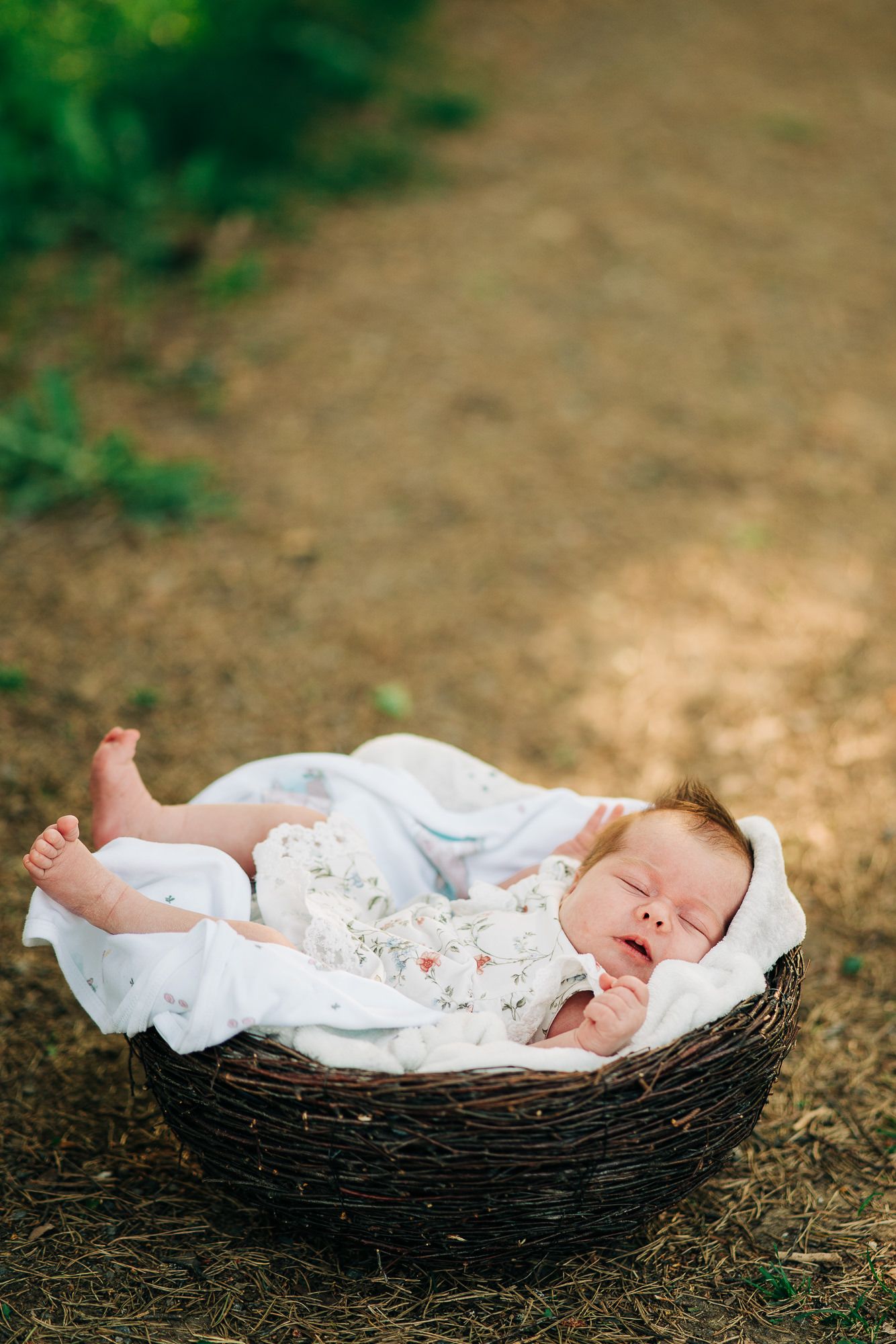 A sleeping baby rests peacefully in a woven basket outdoors, wearing white lace clothing with bare feet.