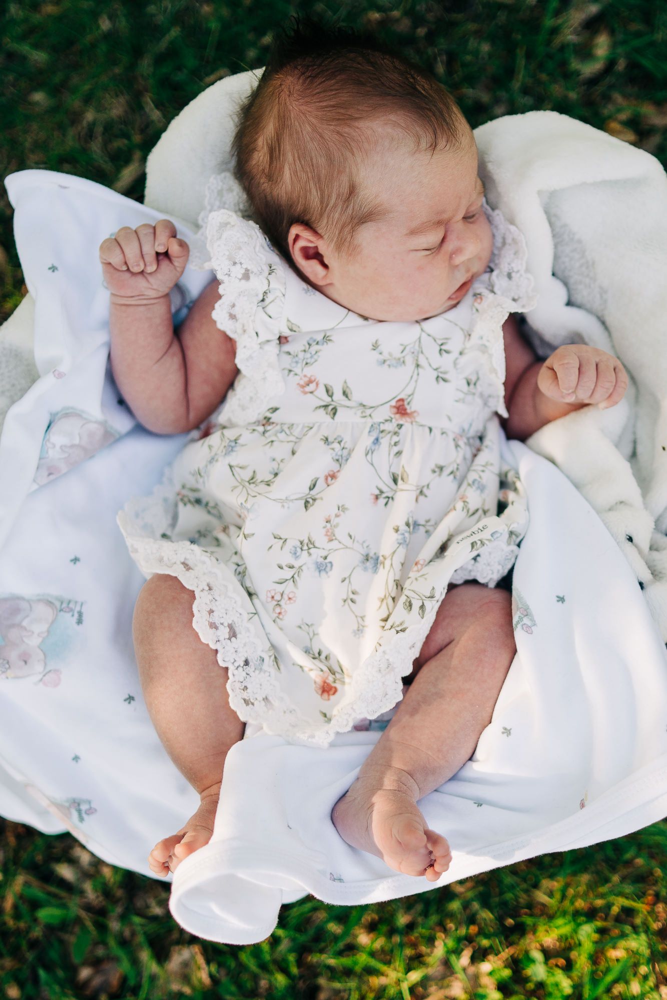 Sleeping newborn lies on white blanket wearing delicate floral romper with lace trim in outdoor setting.
