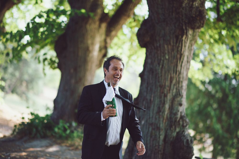 Person in formal black suit laughs while holding beverage outdoors.