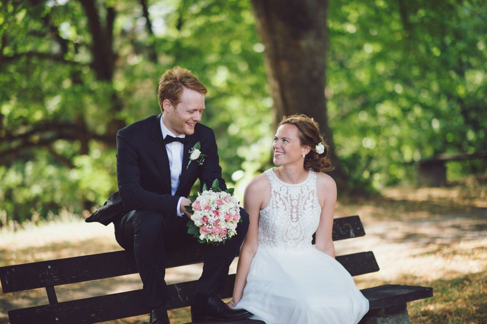 Newlyweds share a joyful moment on a park bench surrounded by lush green trees.