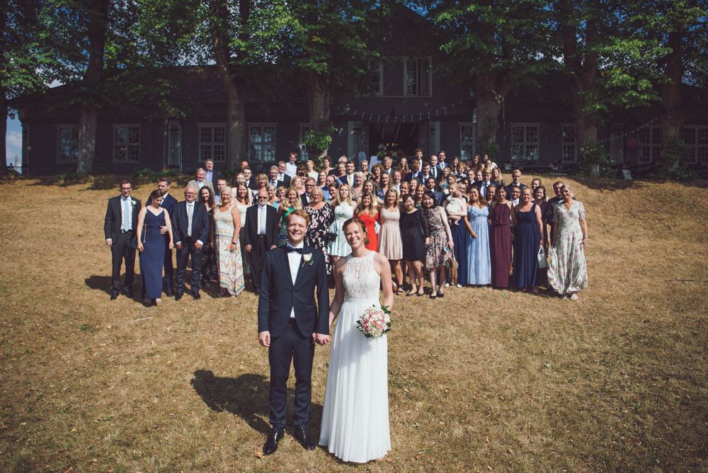 Wedding party and guests gather for large group photo on lawn outside historic building.
