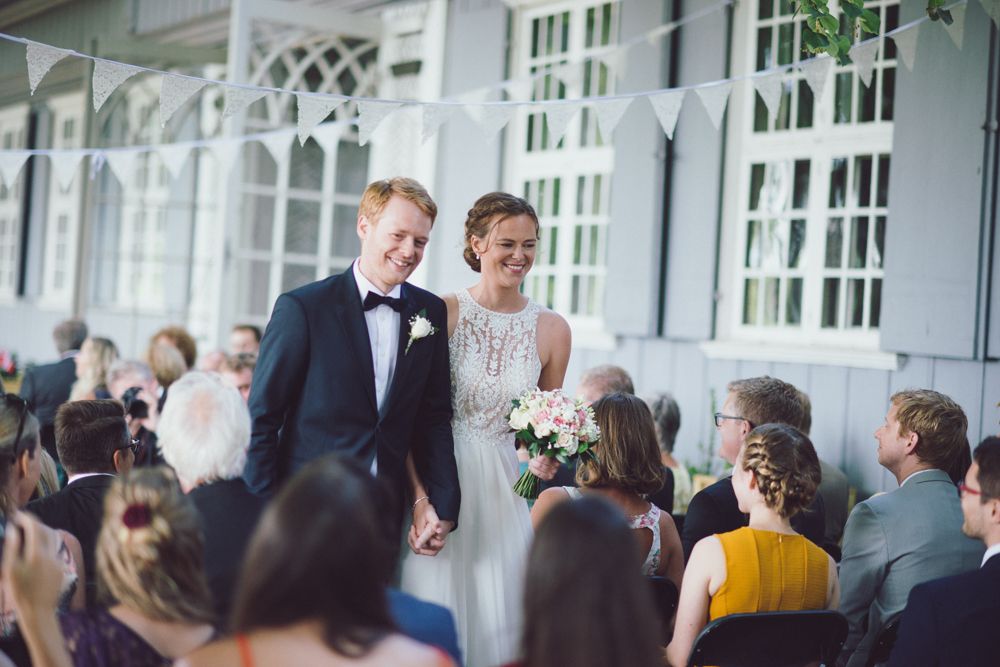 Wedding couple walks down aisle while guests cheer during outdoor ceremony at elegant venue with white columns.