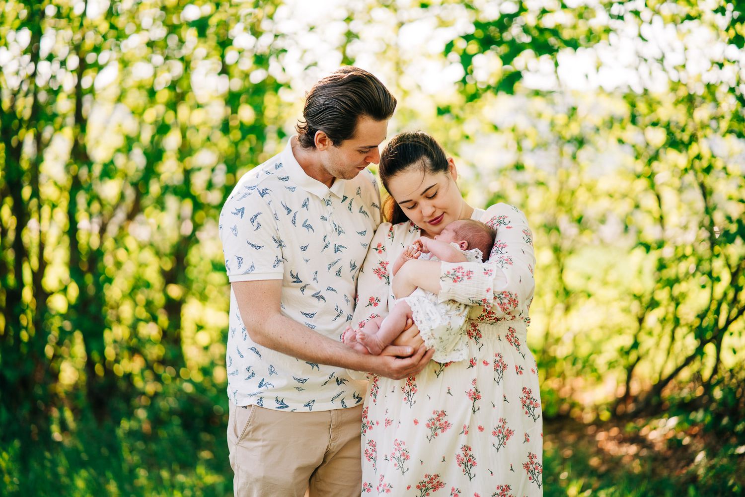 A couple in floral prints holding their newborn baby while standing in a sunlit garden setting with green foliage.