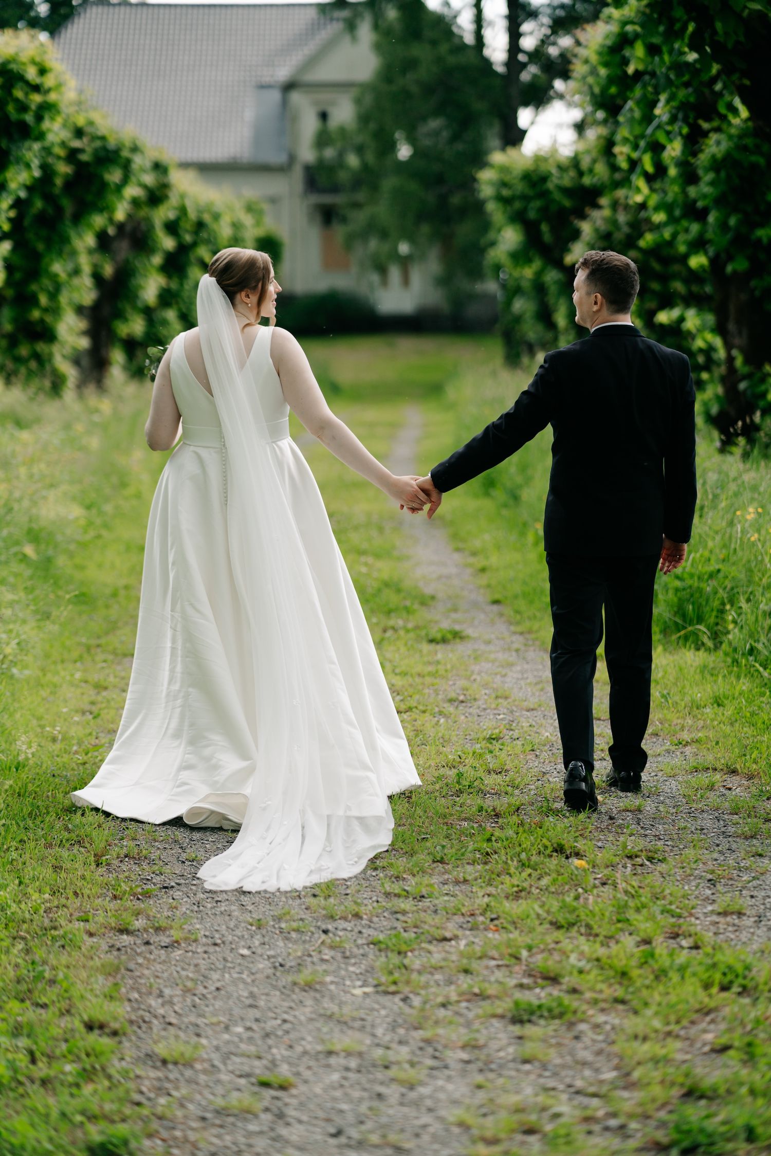 Bride in white gown and groom in black suit walk hand in hand down a gravel path surrounded by lush greenery.