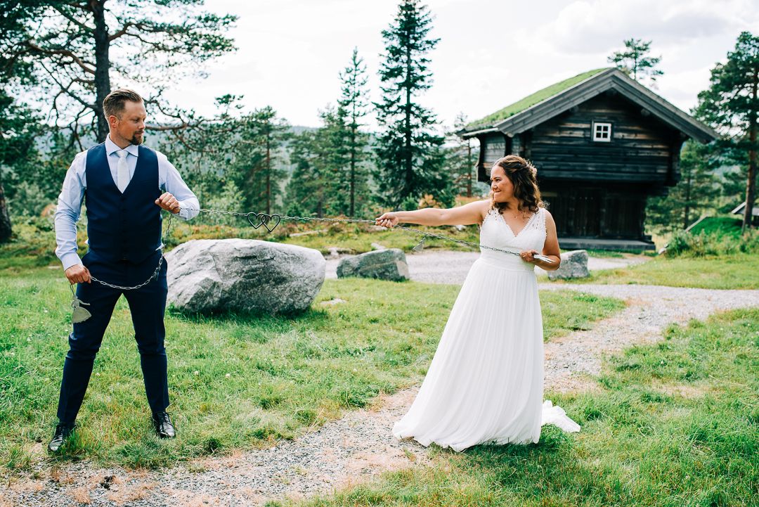 A playful wedding photo of a couple in front of a rustic cabin surrounded by mountain greenery.