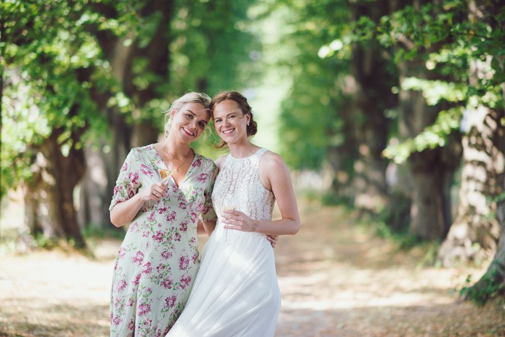 Two women laugh together on tree-lined path during wedding celebration.