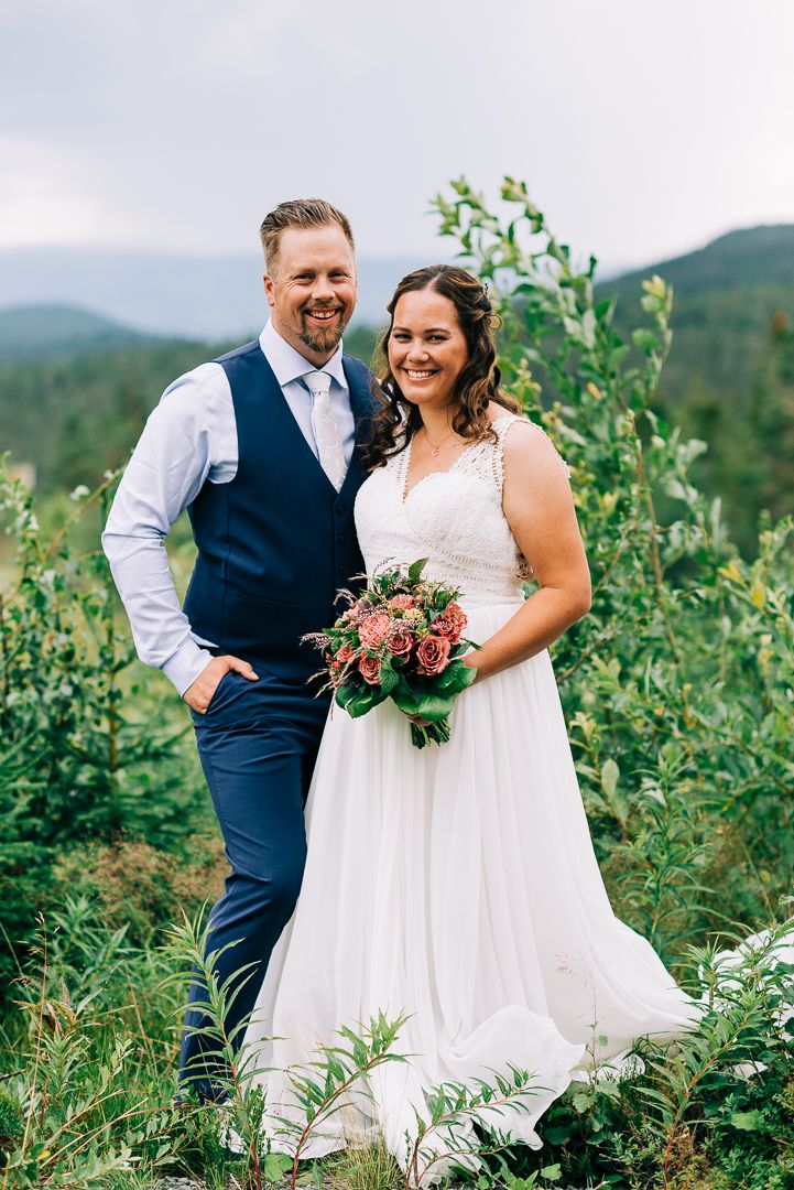 A wedding couple poses on a mountainside with the bride holding a pink bouquet against a scenic natural backdrop.