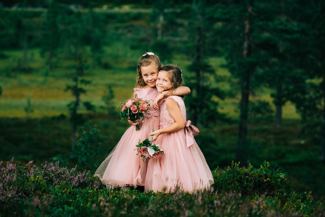 Two flower girls in pink dresses embrace in mountain meadow setting.