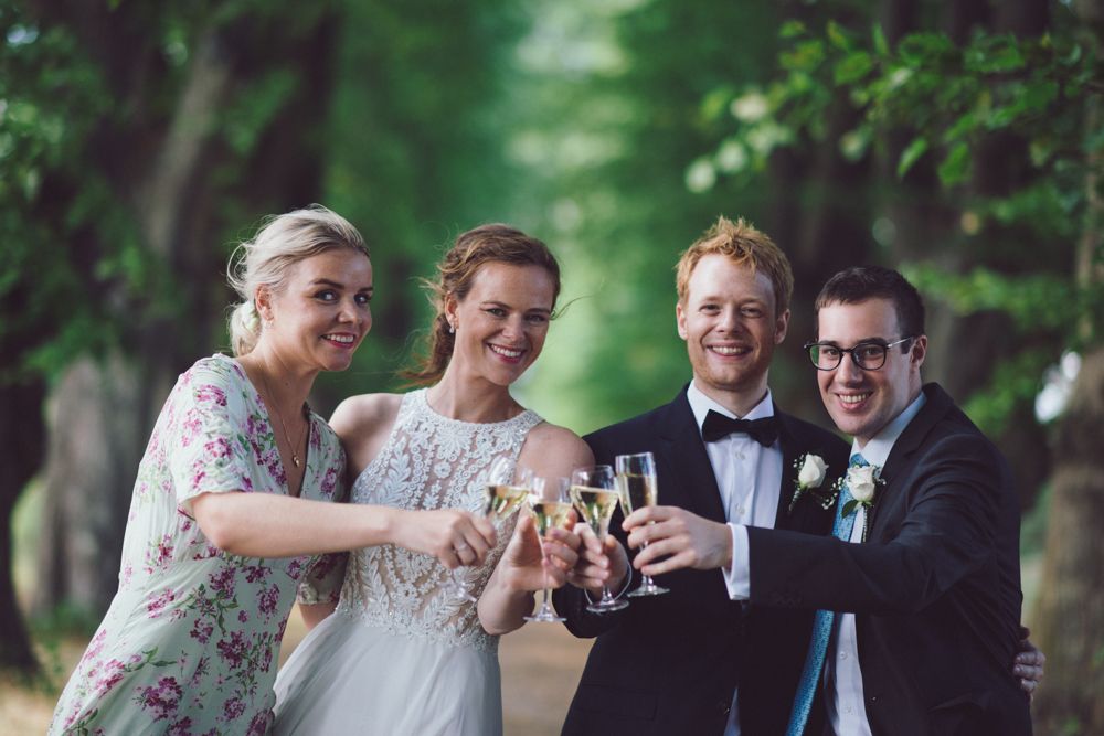 Wedding party toasts with drinks while laughing together on garden path.