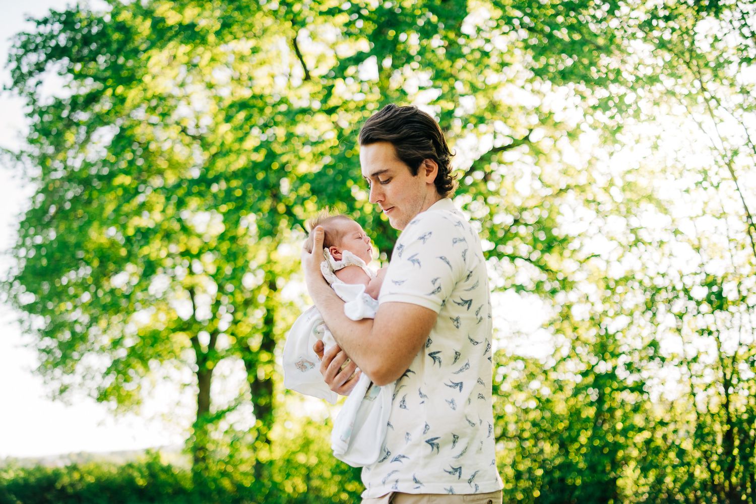 Someone wearing a white floral shirt holds a baby outdoors in a sunlit park setting with green trees and foliage.