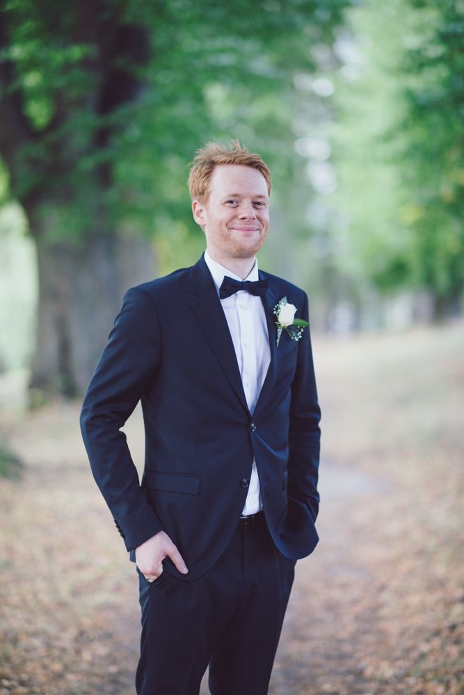 Classic formal portrait of a groom in black tuxedo standing in a tree-lined path.