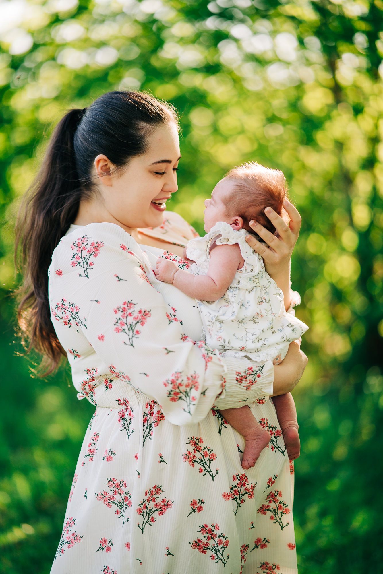 A mother wearing a floral dress holds and smiles at her newborn baby against a soft green outdoor background.