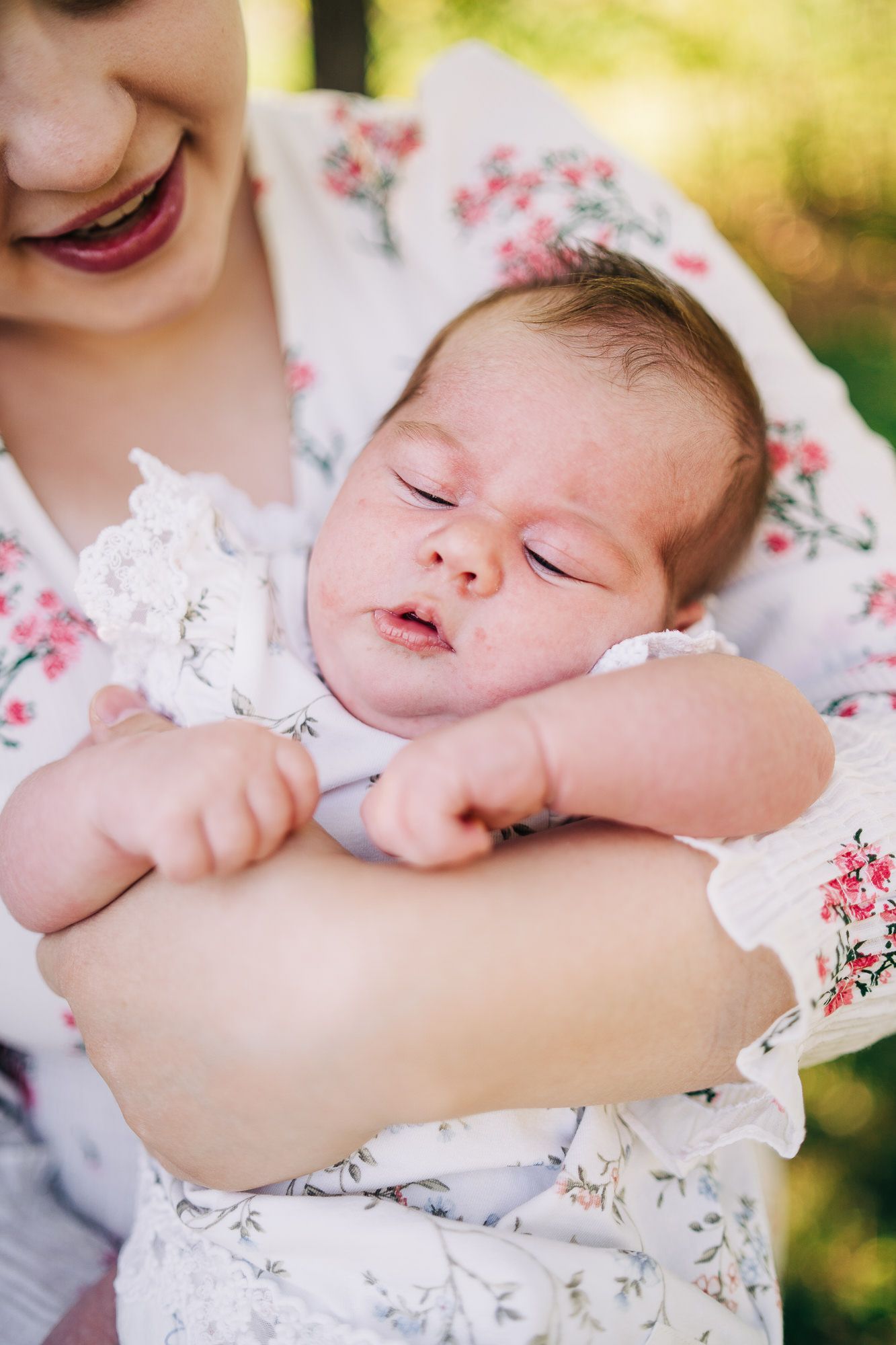Sweet sleeping newborn baby wearing floral dress rests peacefully in cradled arms outdoors in soft natural light.