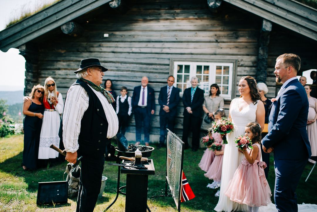 Traditional blacksmith performs demonstration at rustic outdoor Norwegian wedding ceremony near wooden cabin.