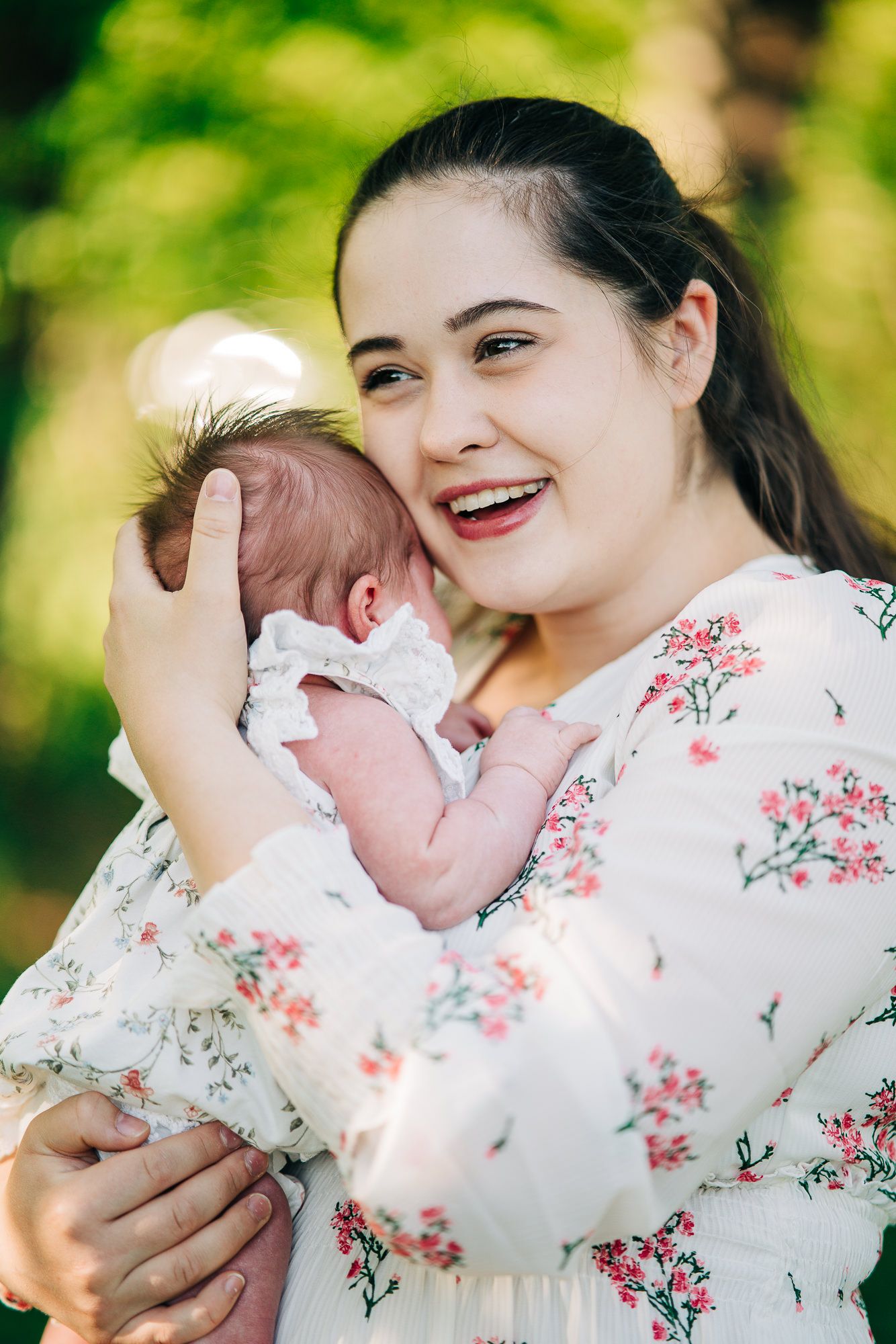 A tender moment between parent and newborn baby outdoors in a floral dress against a green blurred background.
