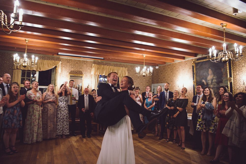 Wedding guests gather around a dance floor in an elegant wood-paneled ballroom with chandeliers.