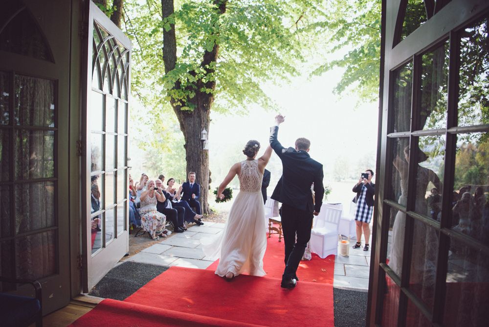 Newlyweds walk down red carpet aisle beneath tree canopy outside historic building.
